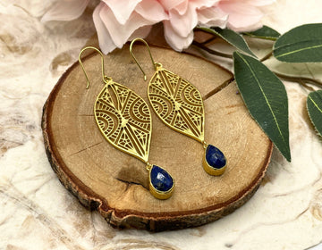 Gold earrings with blue stones on a wooden slice with flowers and leaves.