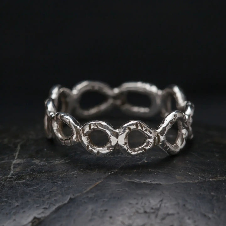 A textured sterling silver ring with a continuous open-link design, resting on a dark, textured stone surface against a black background.