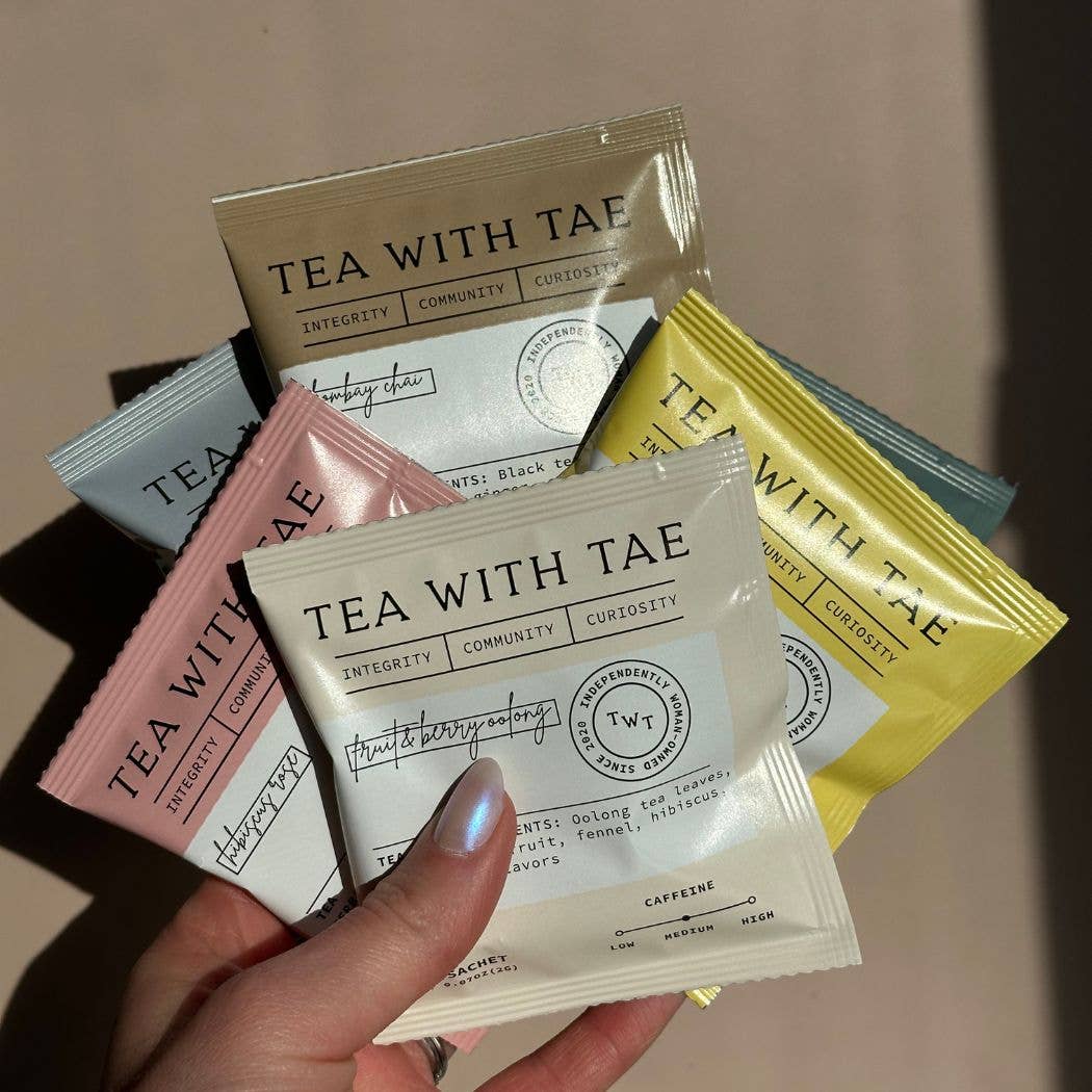 Hand holding multiple 'Tea with Tae' tea bags against a neutral background
