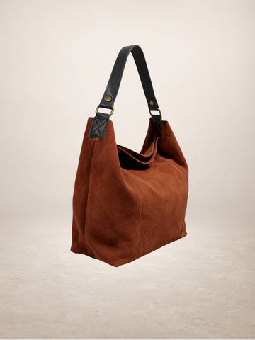 A brown suede hobo-style shoulder bag with black leather straps and gold-tone hardware, shown against a neutral beige studio background.