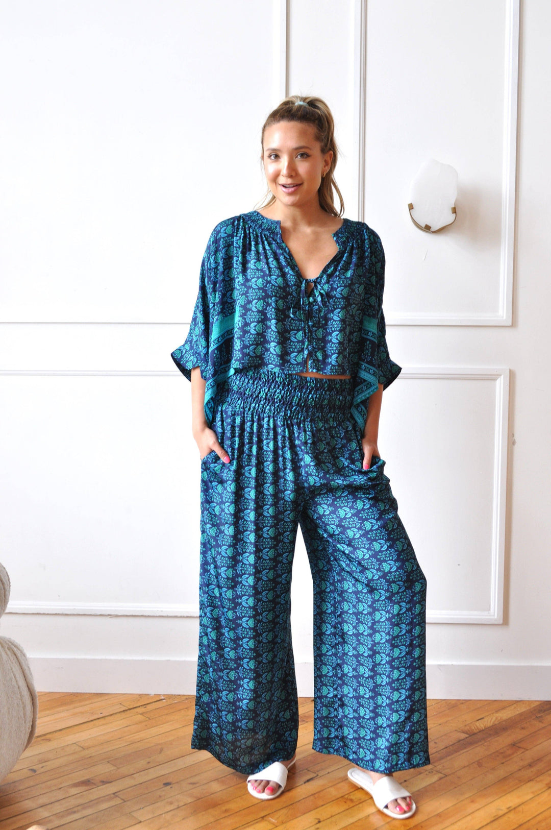 A woman modeling a blue and teal patterned jumpsuit with three-quarter sleeves and wide legs, standing on a wooden floor against a white paneled wall.