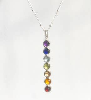 A silver chakra necklace featuring seven vertically aligned round gemstones in purple, blue, green, yellow, orange, brown, and red, suspended from a delicate silver chain against a plain white background.