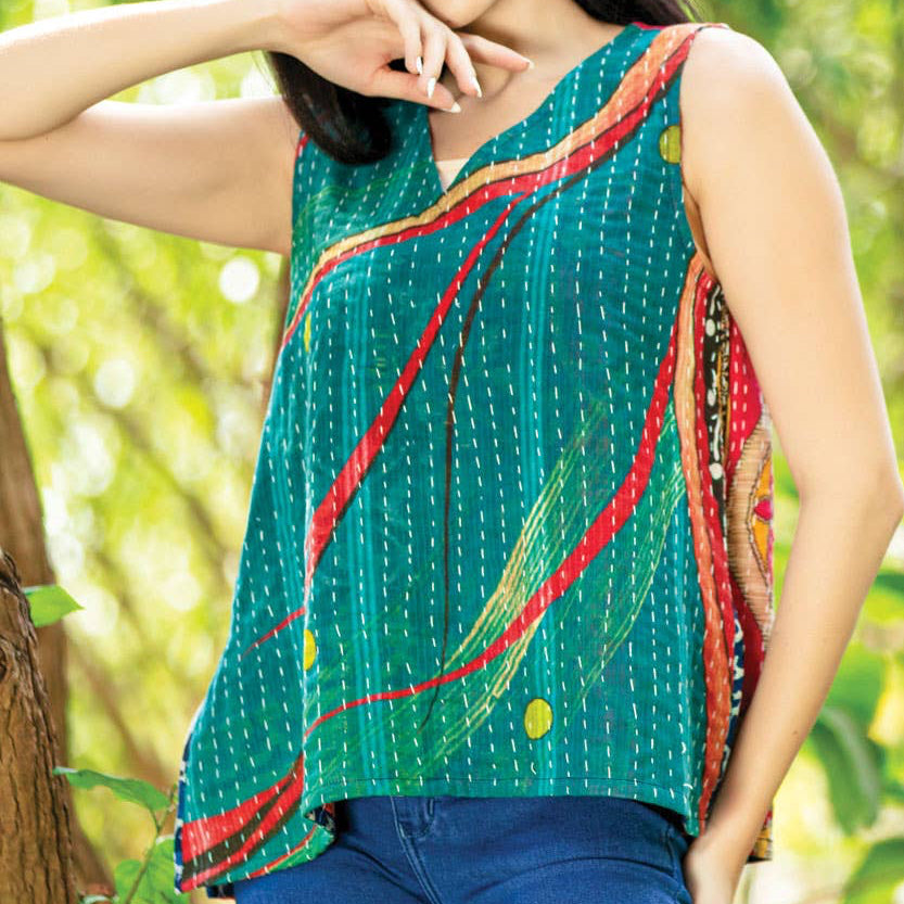 Woman wearing a green sleeveless top with red and yellow patterns, standing outdoors among trees.