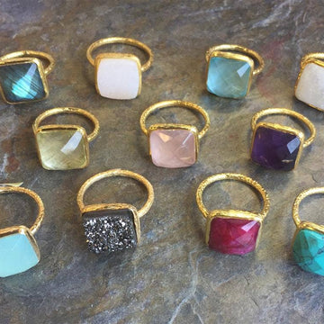 Collection of colorful gemstone rings with gold bands on a stone surface