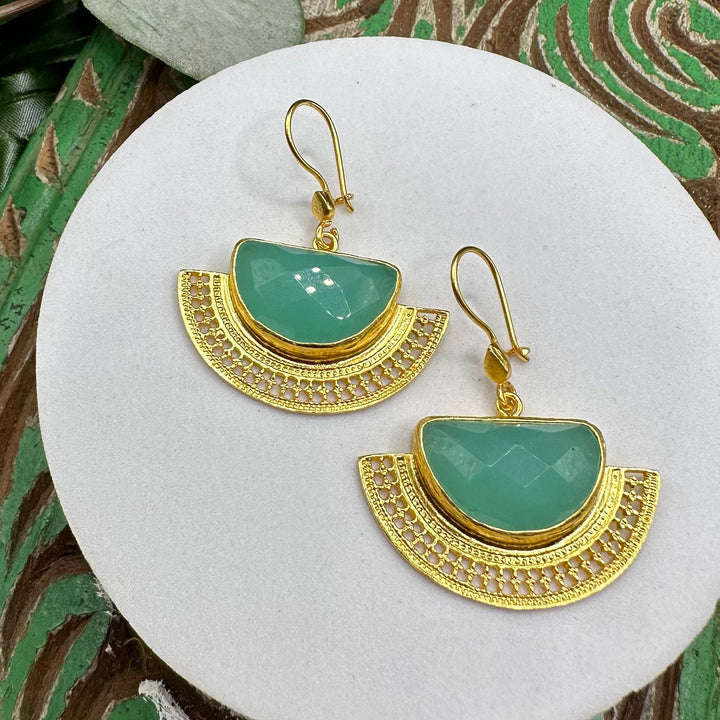 Gold earrings with green semi-precious stones on a white stone surface.