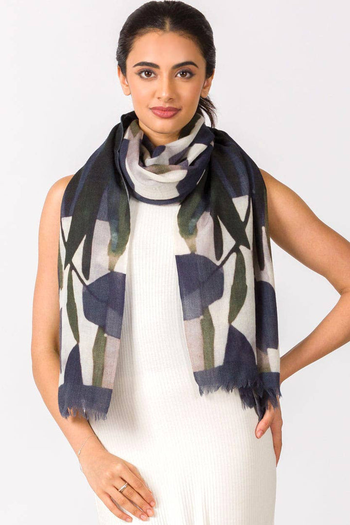 A woman wearing a navy blue and olive green abstract patterned scarf draped around her neck over a white ribbed dress, photographed against a light gray studio background.