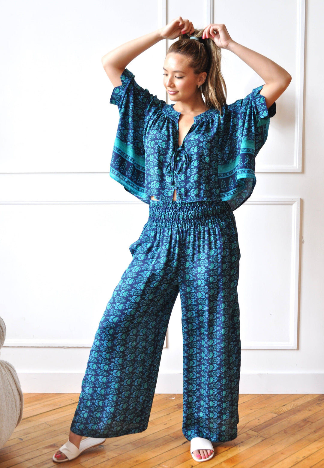 A woman modeling a blue patterned jumpsuit with wide legs and flutter sleeves, standing on a wooden floor against a white paneled wall.