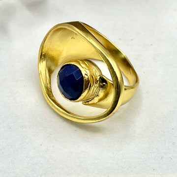 Gold ring with a blue gemstone on a white surface with eucalyptus leaves and driftwood.
