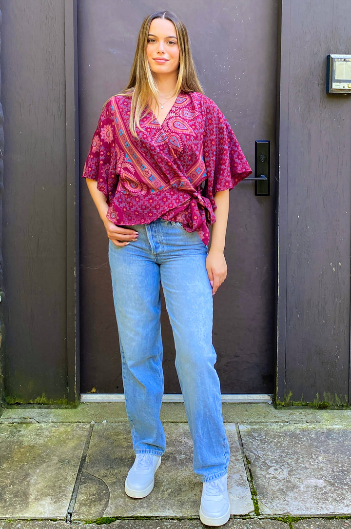 A woman stands in front of a dark door wearing a bright pink and purple paisley wrap blouse, light blue jeans, and white sneakers.
