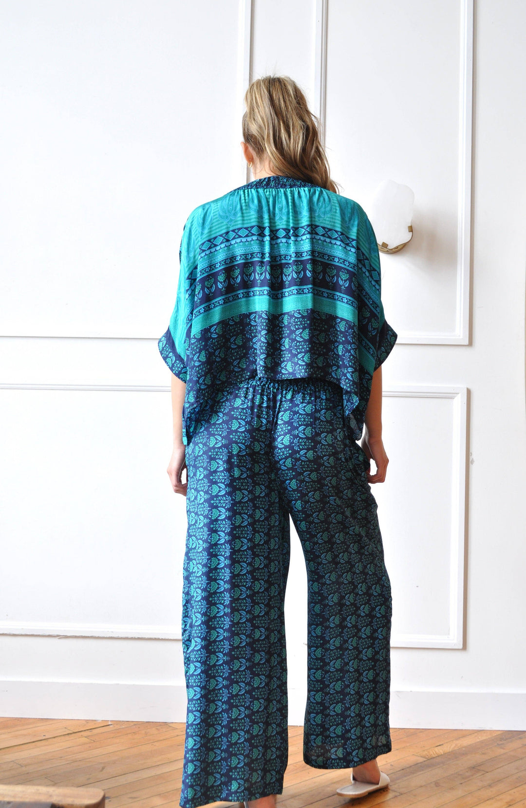 A woman viewed from behind wearing a matching two-piece lounge set with a teal and dark blue patterned kaftan top and wide-leg pants, standing on a wooden floor against a white paneled wall.