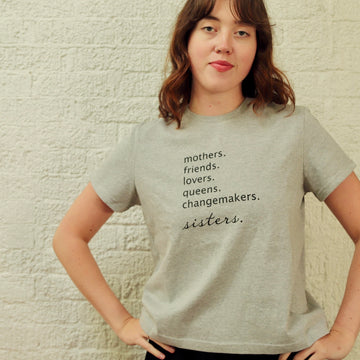 A woman wearing an oversized heather grey cropped t-shirt with the text 'mothers. friends. lovers. queens. changemakers. sisters'