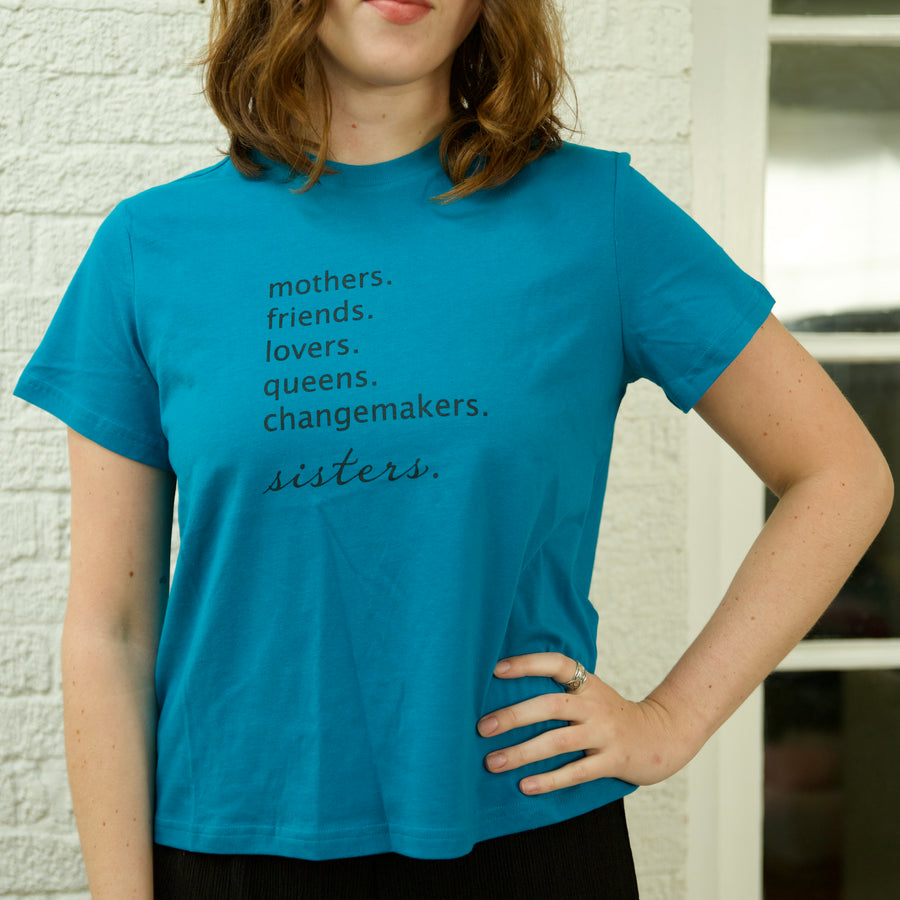 A person wearing an oversized teal cropped t-shirt with the text 'mothers. friends. lovers. queens. changemakers. sisters.'