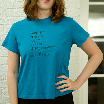A person wearing an oversized teal cropped t-shirt with the text 'mothers. friends. lovers. queens. changemakers. sisters.'