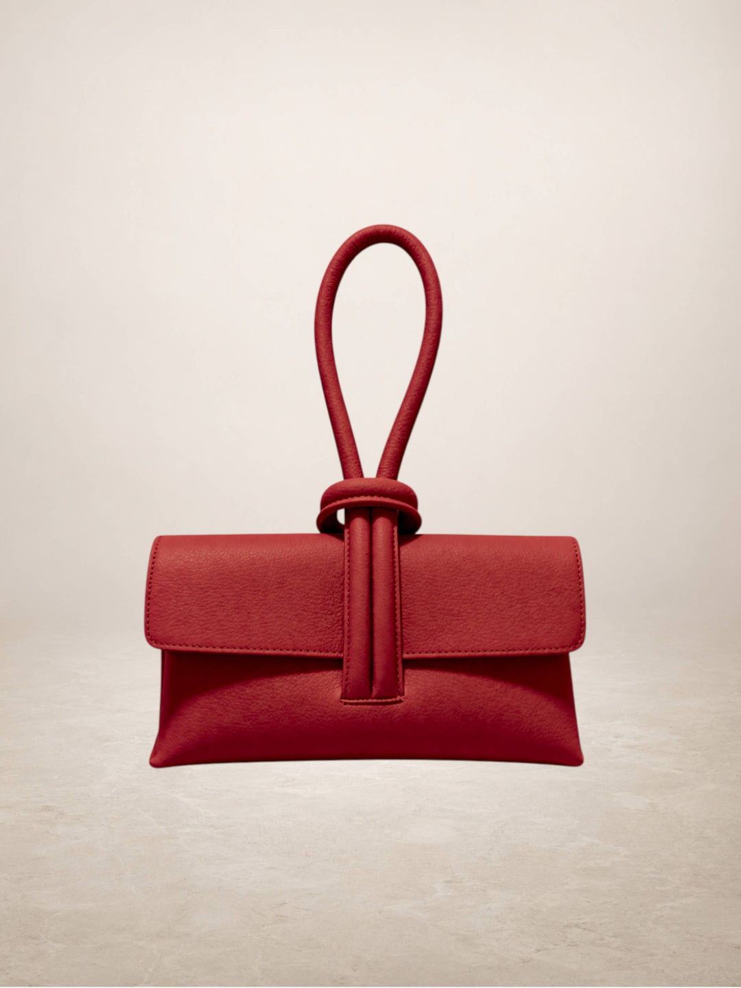 A structured red leather clutch handbag with a top loop handle knotted in front of the flap, shown on a neutral beige background.