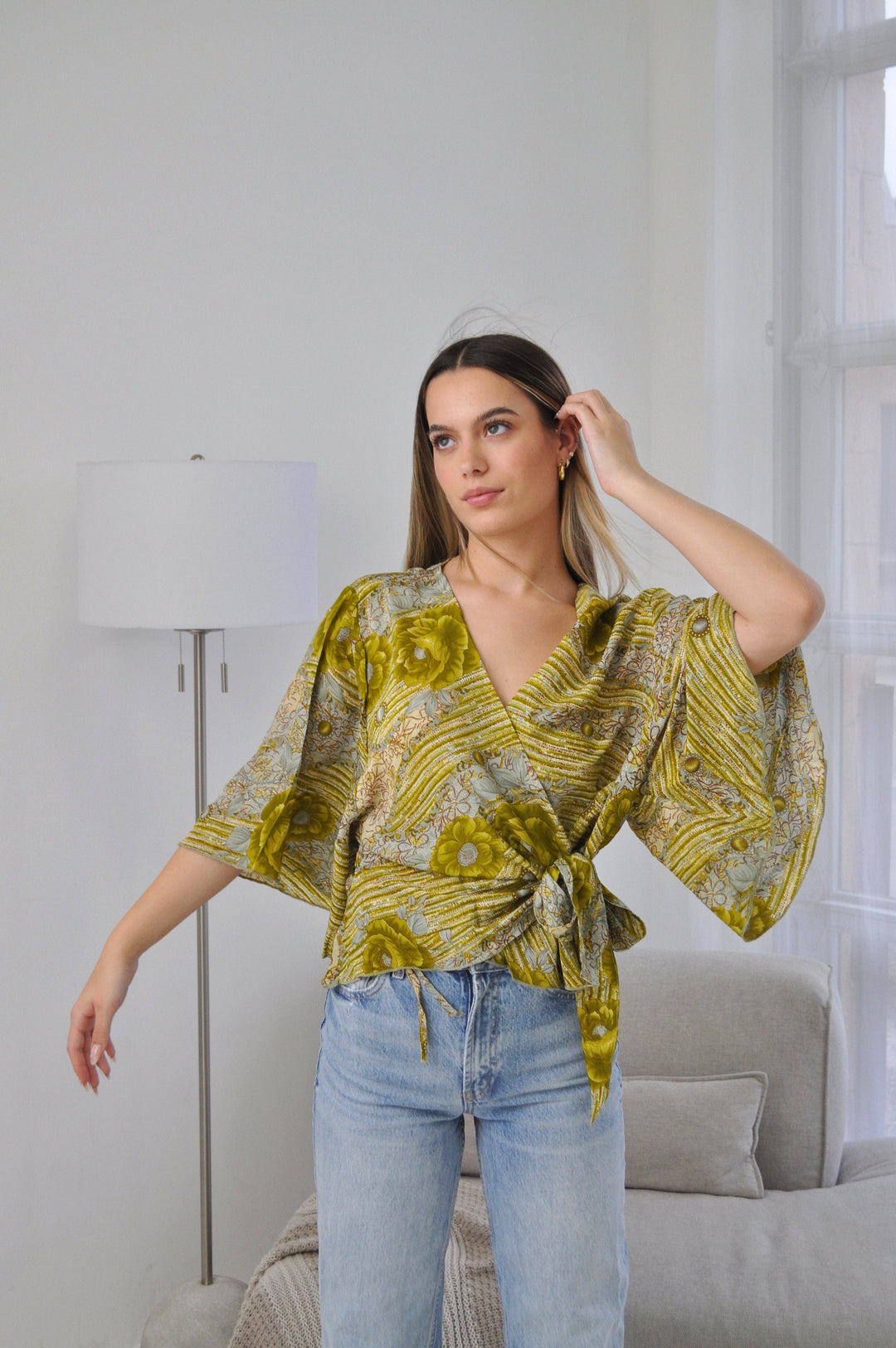 A woman modeling a yellow and green patterned wrap top with wide bell sleeves, paired with light blue jeans, standing in a bright, modern room with a floor lamp and sofa.