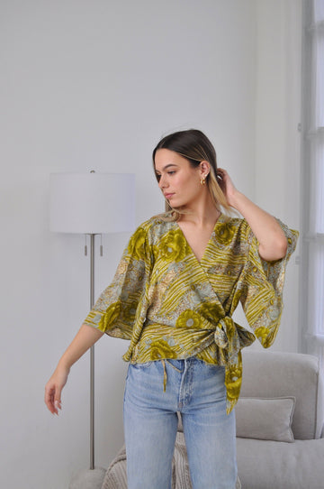 A woman wearing a yellow and green patterned wrap top with wide sleeves and a tie waist, paired with light blue jeans, standing in a bright room with a floor lamp and sofa.