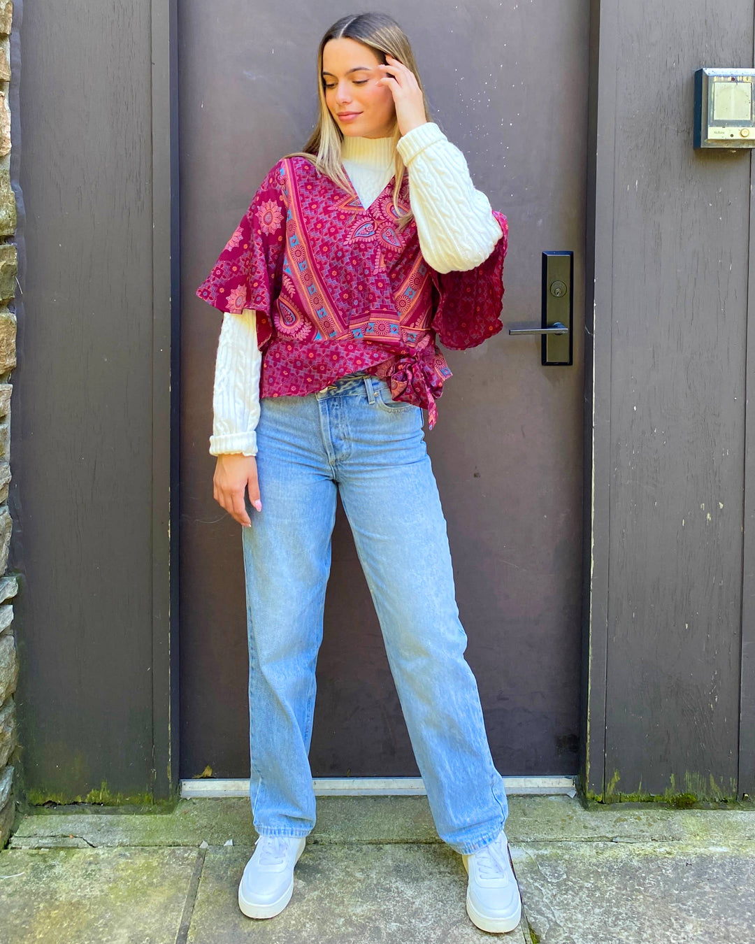 A woman stands in front of a dark brown wooden door wearing a bright pink and red paisley patterned top with white ribbed knit sleeves, light blue jeans, and white sneakers.