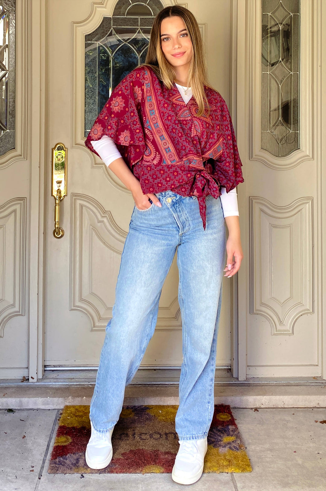 A woman stands in front of a beige paneled door wearing a red and blue paisley patterned poncho over a white shirt, light-wash blue jeans, and white sneakers.