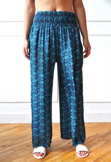 A person wearing loose, wide-leg pants with a blue and teal geometric pattern, paired with white slide sandals, standing on a wooden floor against a white paneled wall.