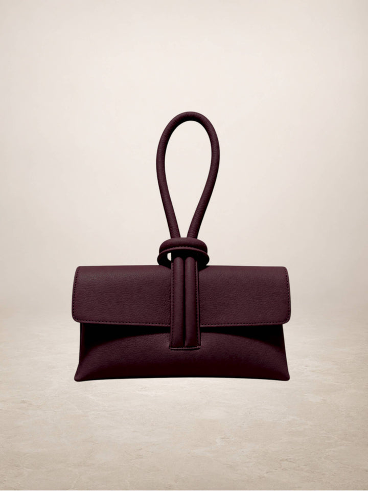 A dark plum purple leather clutch bag with a unique knotted top handle, displayed against a neutral beige background.