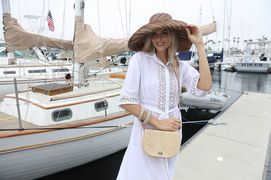 Woman in a white dress and large hat standing on a dock with boats in the background