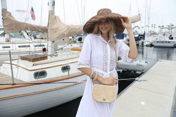 Woman in a white dress and large hat standing on a dock with boats in the background