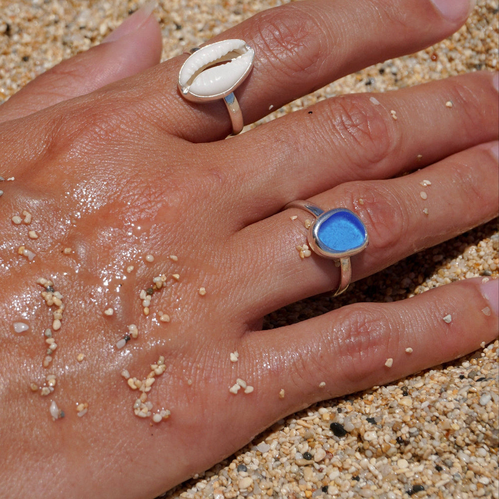 Hand with two rings, one featuring a cowrie shell design and the other with a blue stone, on a sandy background.