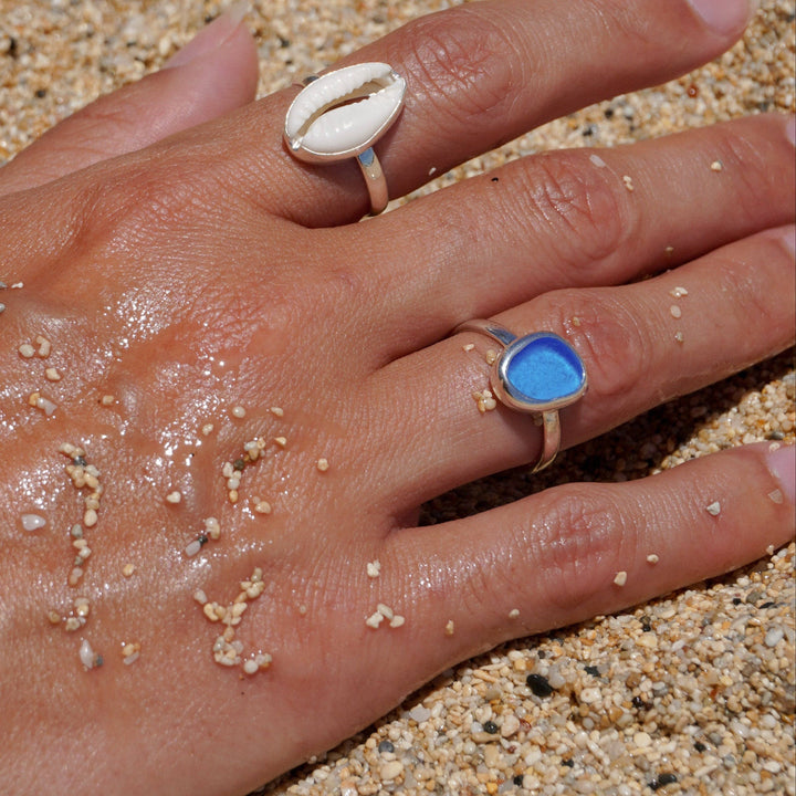 Hand with two rings, one featuring a cowrie shell design and the other with a blue stone, on a sandy background.