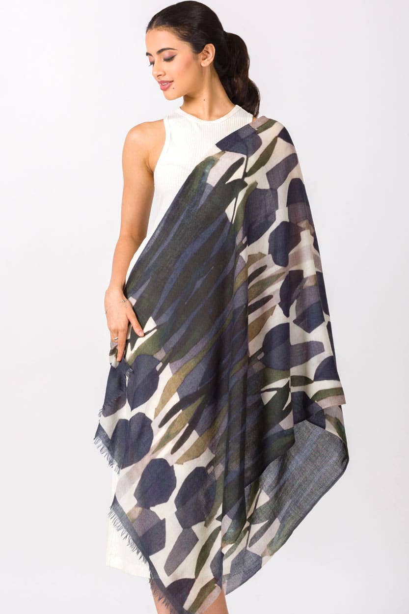 A woman modeling a lightweight draped scarf with an abstract floral pattern in navy blue, olive green, and beige, paired with a white sleeveless top against a plain white studio background.